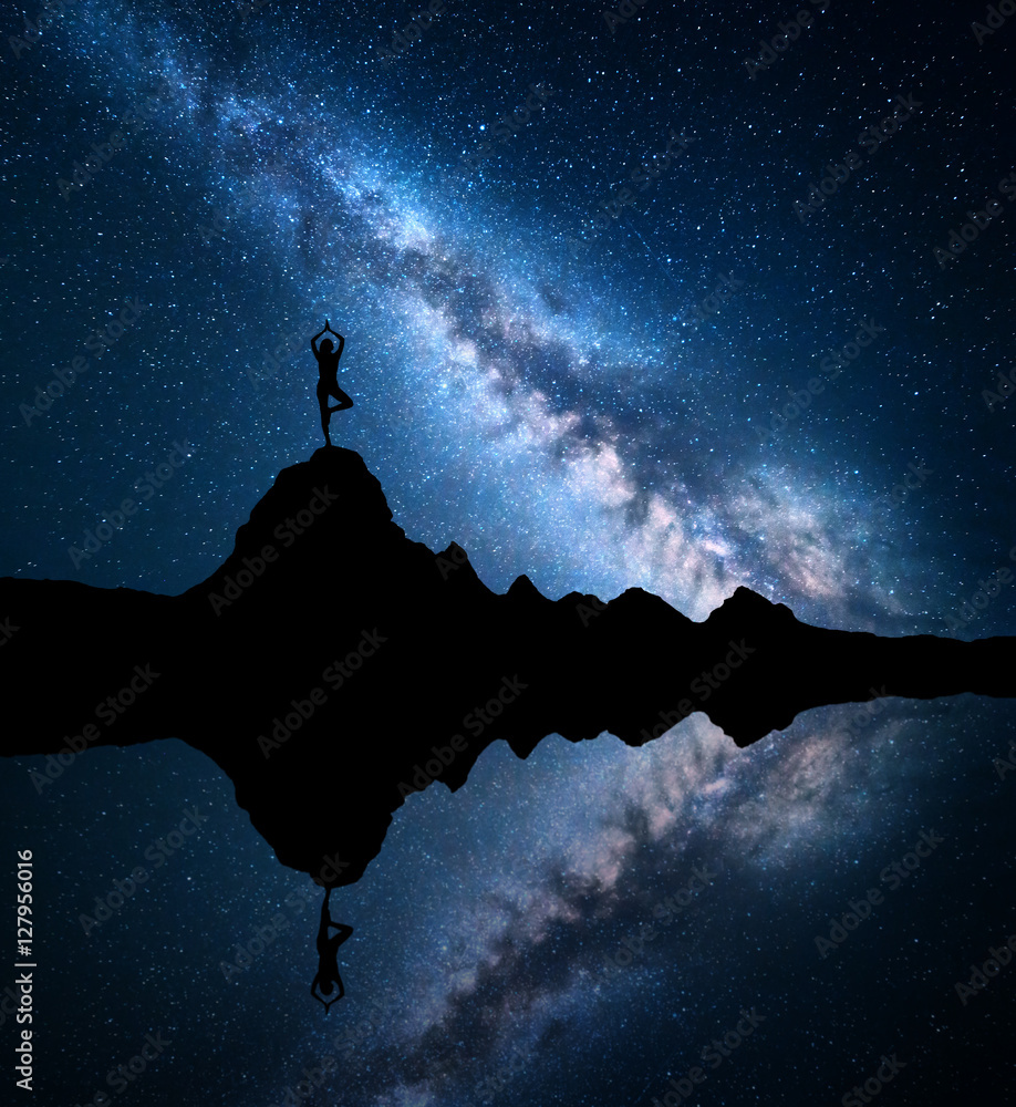 银河系。一个站着的女人和sk在湖边的山峰上练习瑜伽的剪影