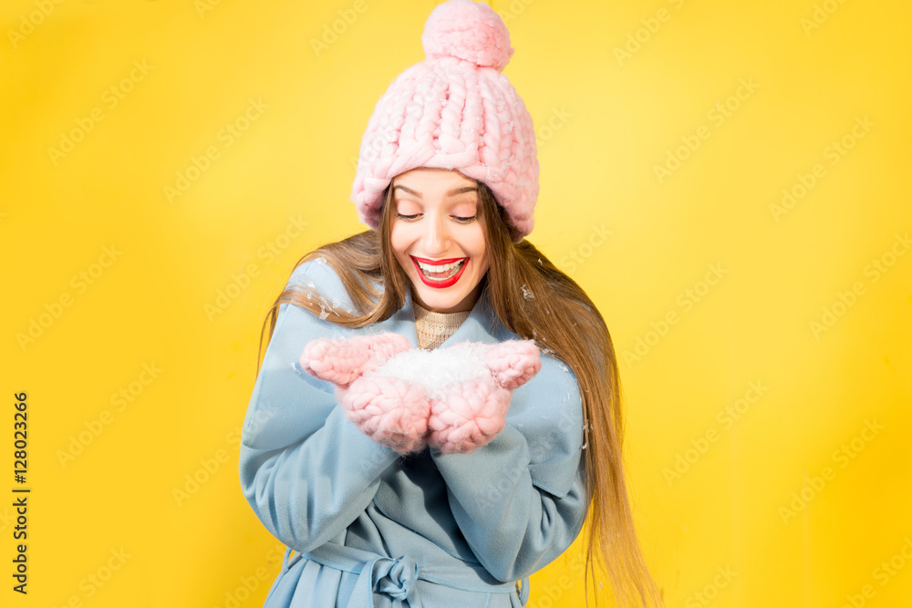戴着针织帽子和手套的年轻女子站在黄色的墙上吹雪。Colorfu