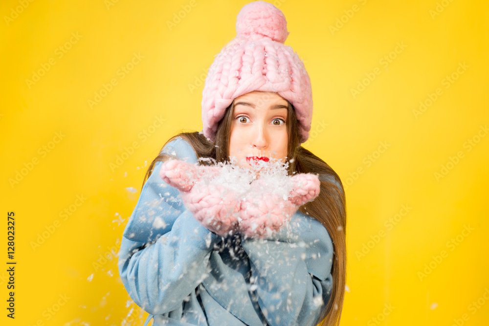 戴着针织帽子和手套的年轻女子站在黄色的墙上吹雪。Colorfu