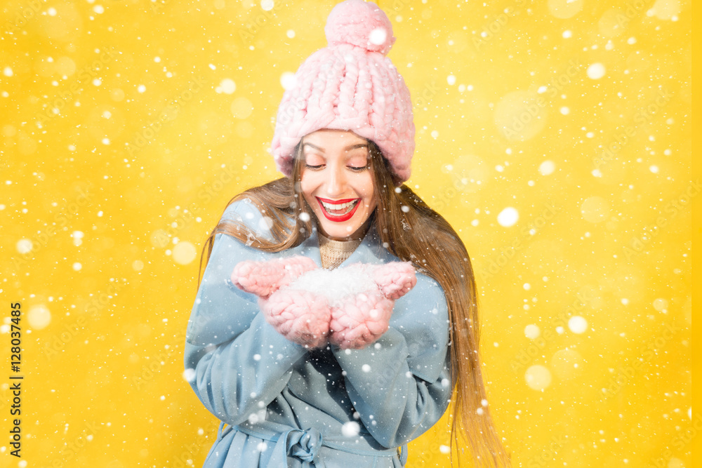 戴着针织帽子和手套的年轻女子在黄色的墙壁背景上抱着雪。五颜六色的幸福w