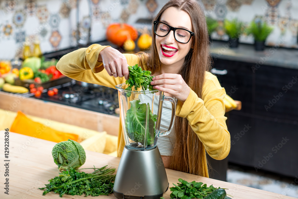 Young smiling woman making smoothie with fresh greens in the blender in kitchen at home. Healthy veg