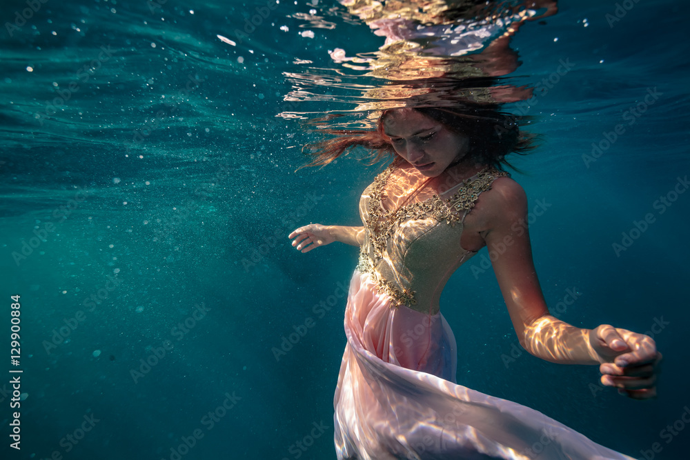 深蓝色海洋背景下，一位身穿金粉色连衣裙的模特在水下