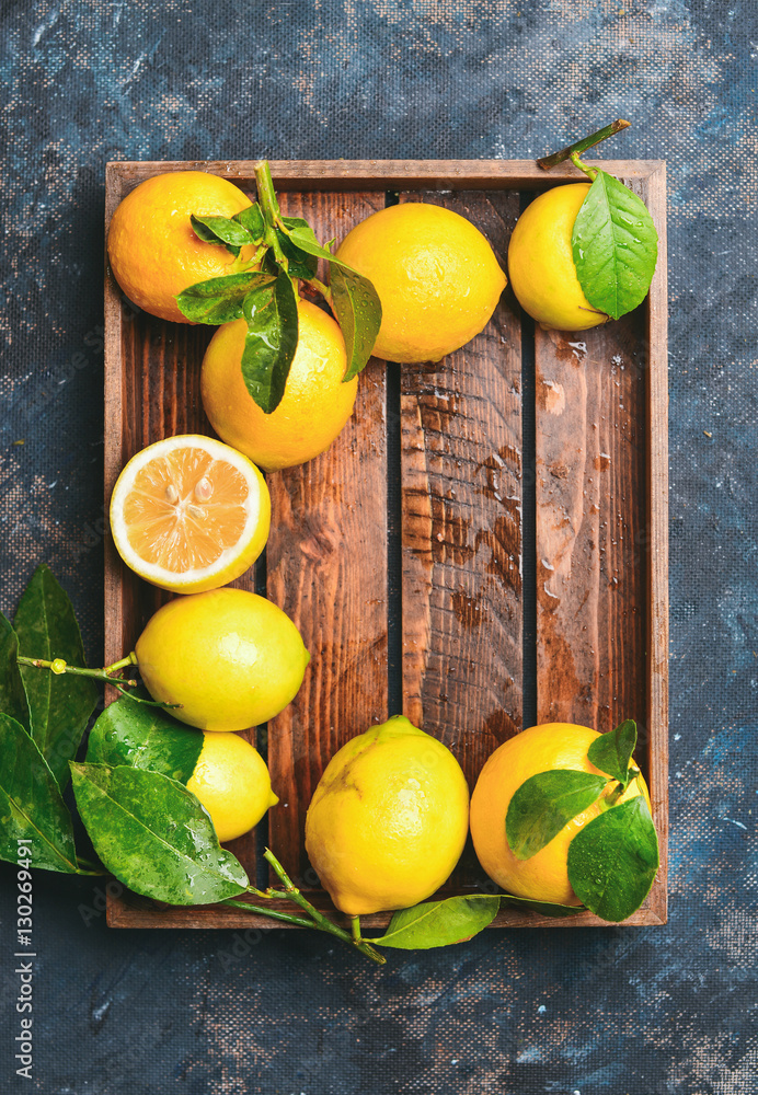Freshly picked lemons with leaves in wooden tray over dark blue shabby plywood background, top view,