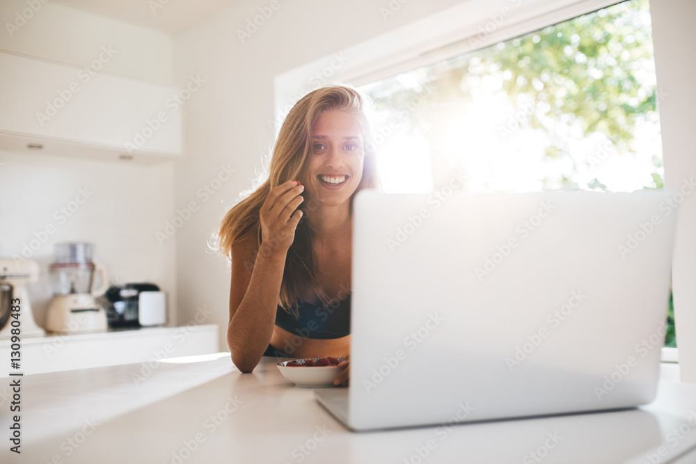 Beautiful girl eating berries and using laptop in kitchen