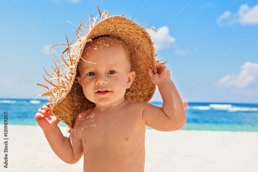 Funny photo of happy baby boy on beach with straw hat and dirty face covered with sand. Family trave