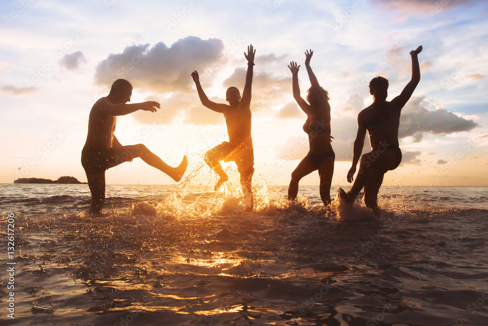 group of happy friends having fun together on the beach at sunset, jumping and dancing with water sp