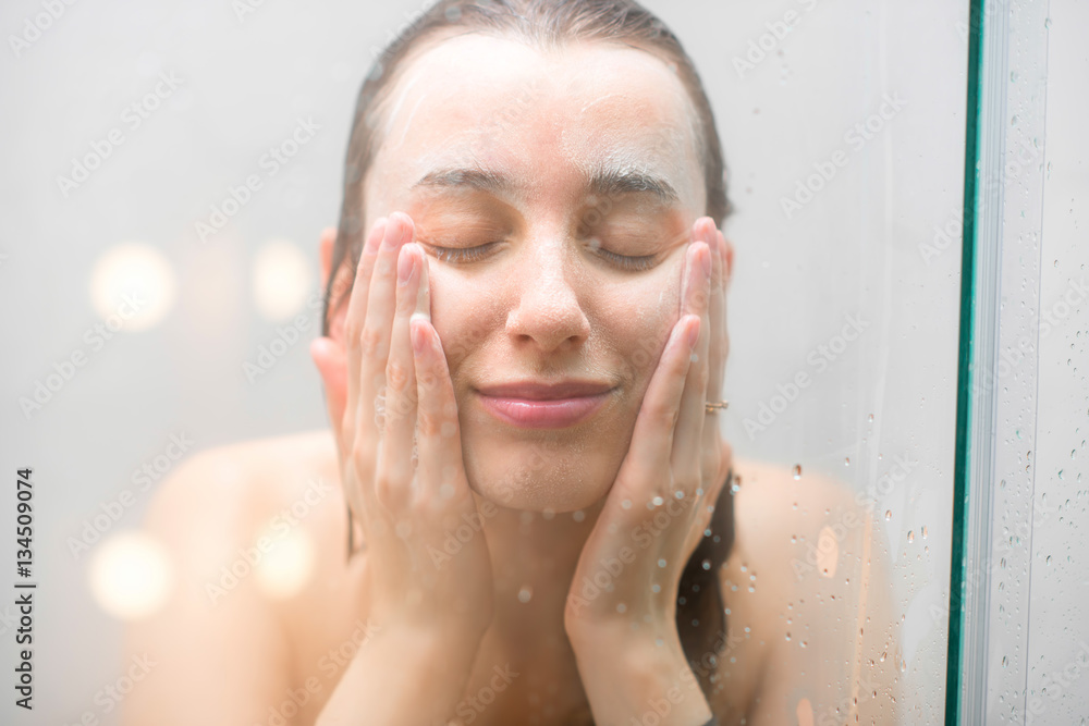淋浴时，一个湿漉漉的脸上涂着肥皂的女人站在玻璃后面的特写。Imag