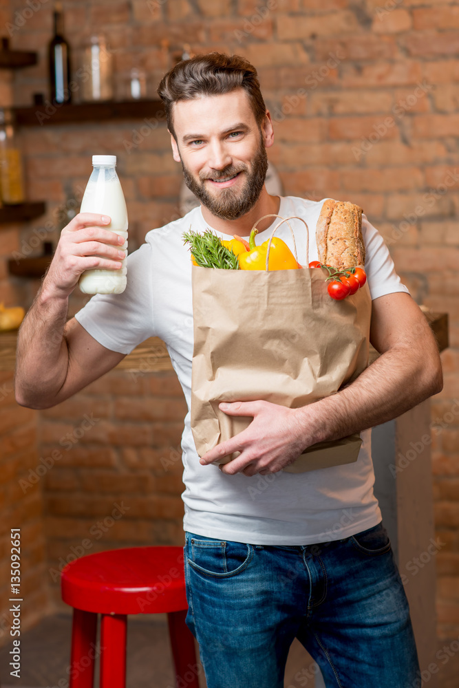 Handsome man holding milk with paper bag full of healthy food in the kitchen