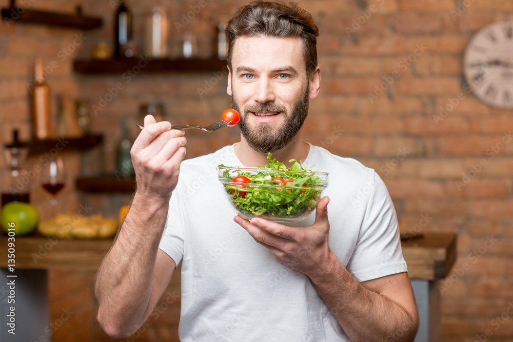 Handsome bearded man in white t-shirt eating salad with tomatoes in the kitchen. Healthy and vegan f