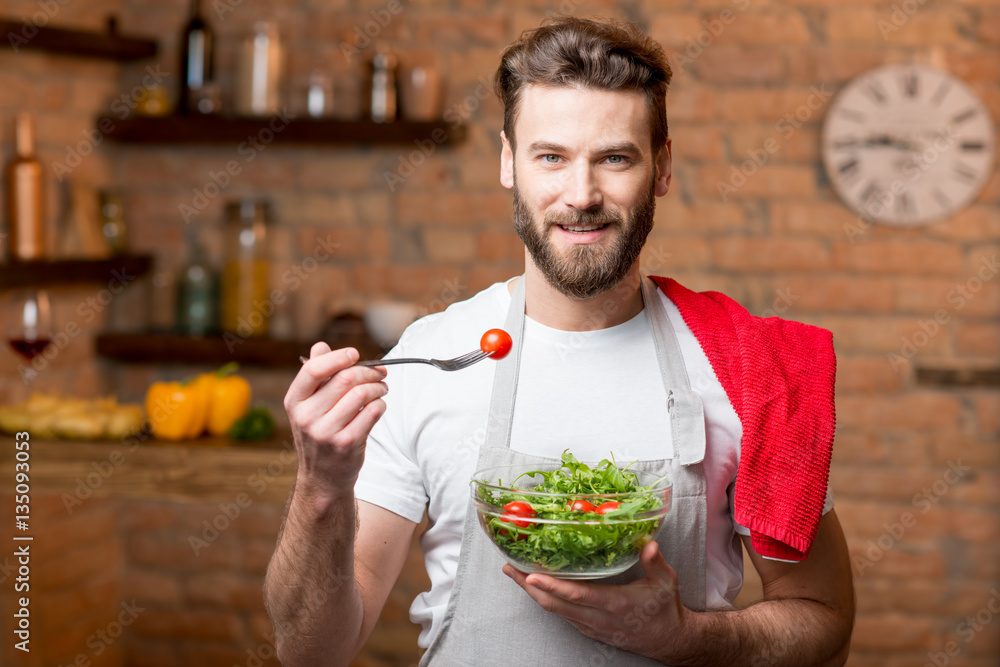 Handsome bearded man in white t-shirt and apron eating salad with tomatoes in the kitchen. Healthy a