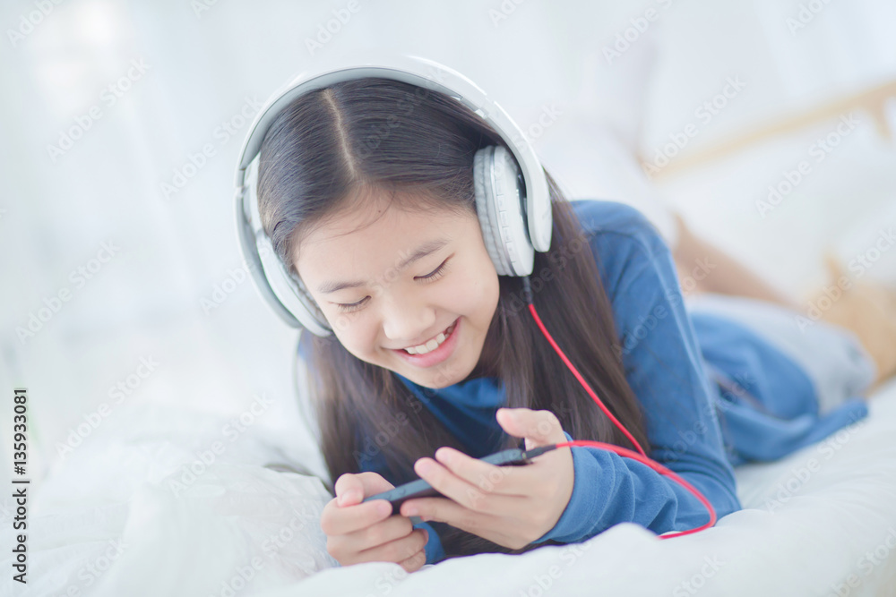 Pretty Asian girl using headphone for listen music by smartphone on the bed in her bedroom