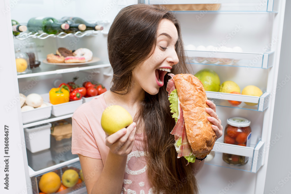 Young woman choosing between apple and big sandwich standing in front of the refrigerator full of ve