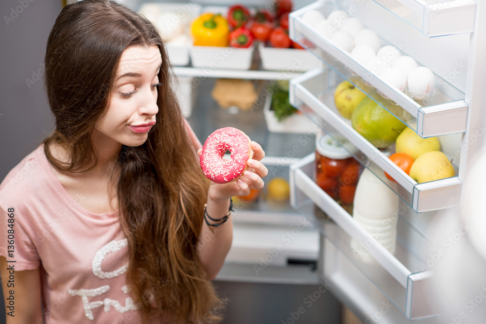Portrait of a young sad woman in the sleepwear with sweet donuts standing near the open refrigerator