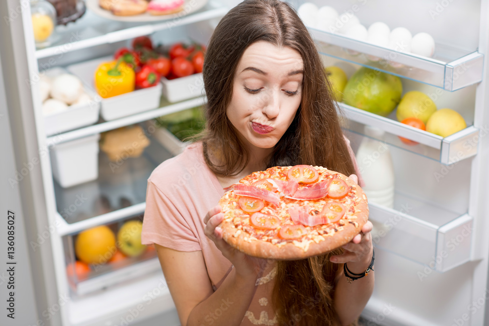 Young woman with pizza standing near the open refrigerator full of fruits and vegetales