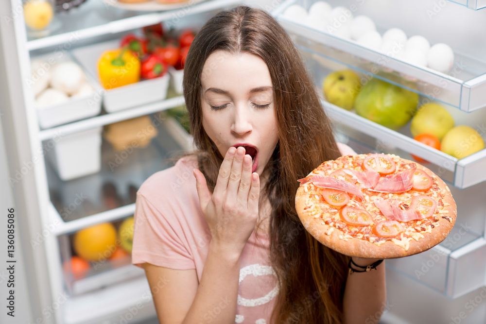Young woman with pizza yawning near the open refrigerator full of fruits and vegetales