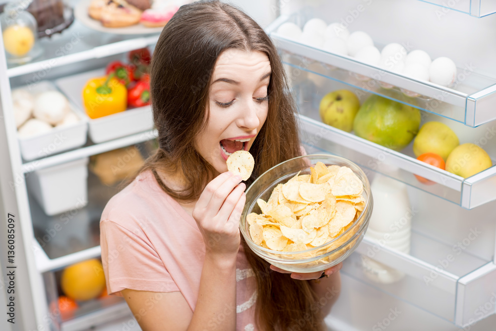 Young woman eating potato chips near the refrigerator full of healthy food