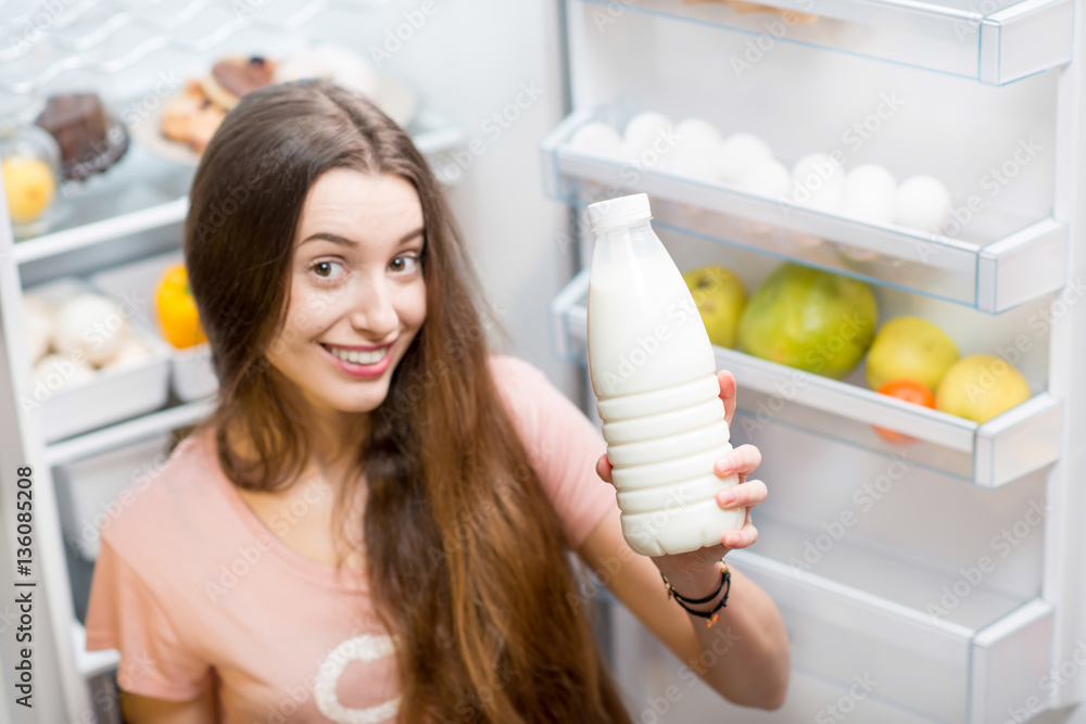Young smiling woman showing bottle of milk standing in front of the refrigerator