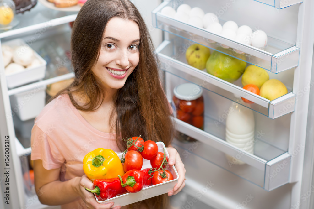Portrait of a young smiling woman with vegetables standing in front of the open refrigerator