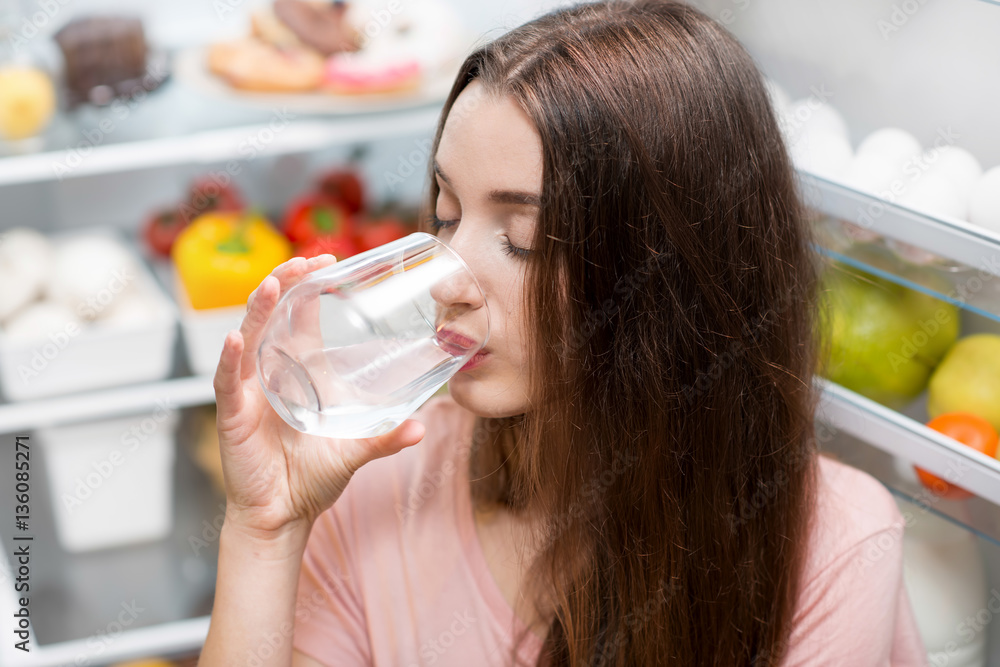 Young woman drinking water near the refrigerator