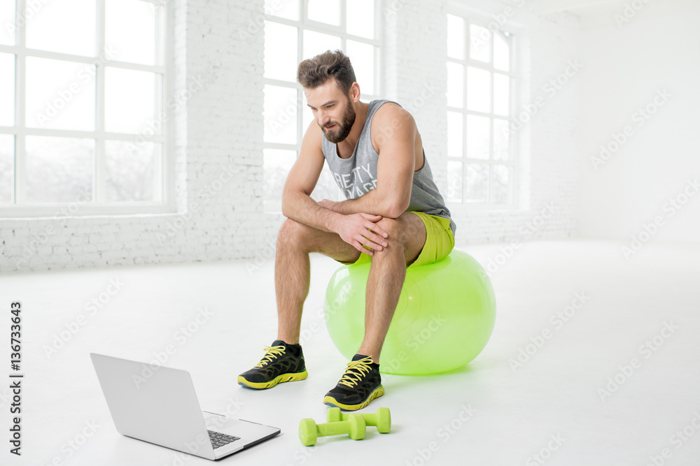 Man watching online trainig with laptop sitting on the fitball in the gym