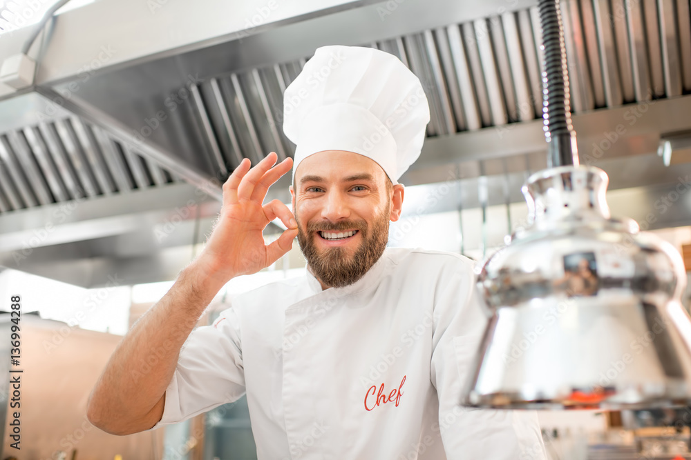 Portrait of a handsome chef cook in uniform showing delicious sign at the restaurant kitchen