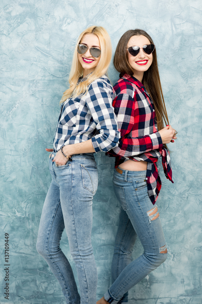 Portrait of two female friends in checkered shirts, jeans and sunglasses on the blue painted wall ba