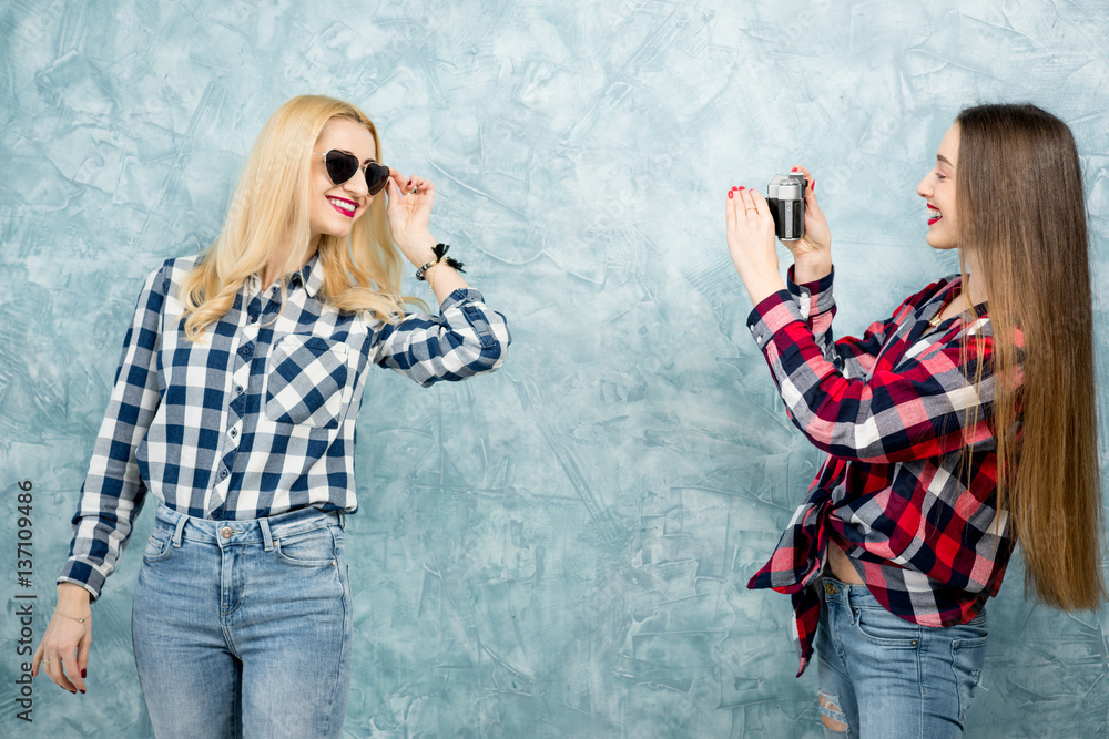 Two female friends in checkered shirts and jeans photographing with retro camera on the blue painted