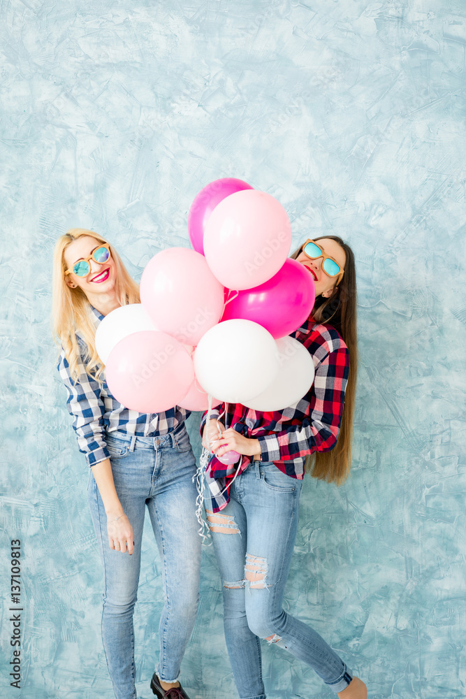 Two female friends in checkered shirts having fun with air balloons on the blue wall background