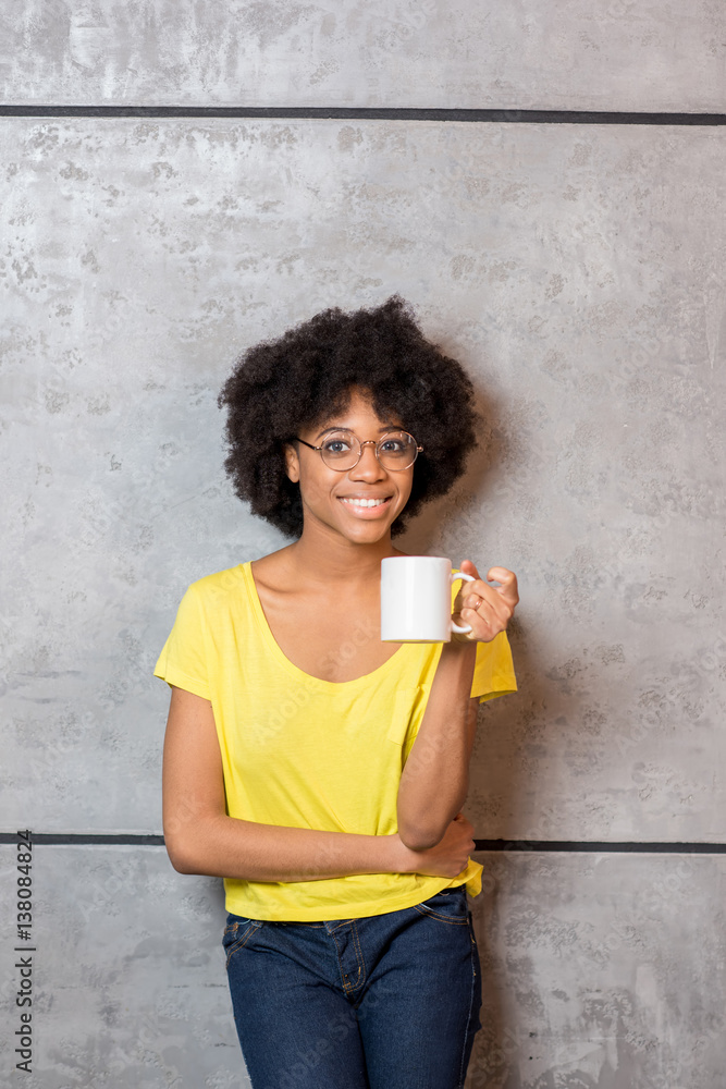 Portrait of a beautiful african woman in yellow t-shirt drinking coffee near the gray wall indoors