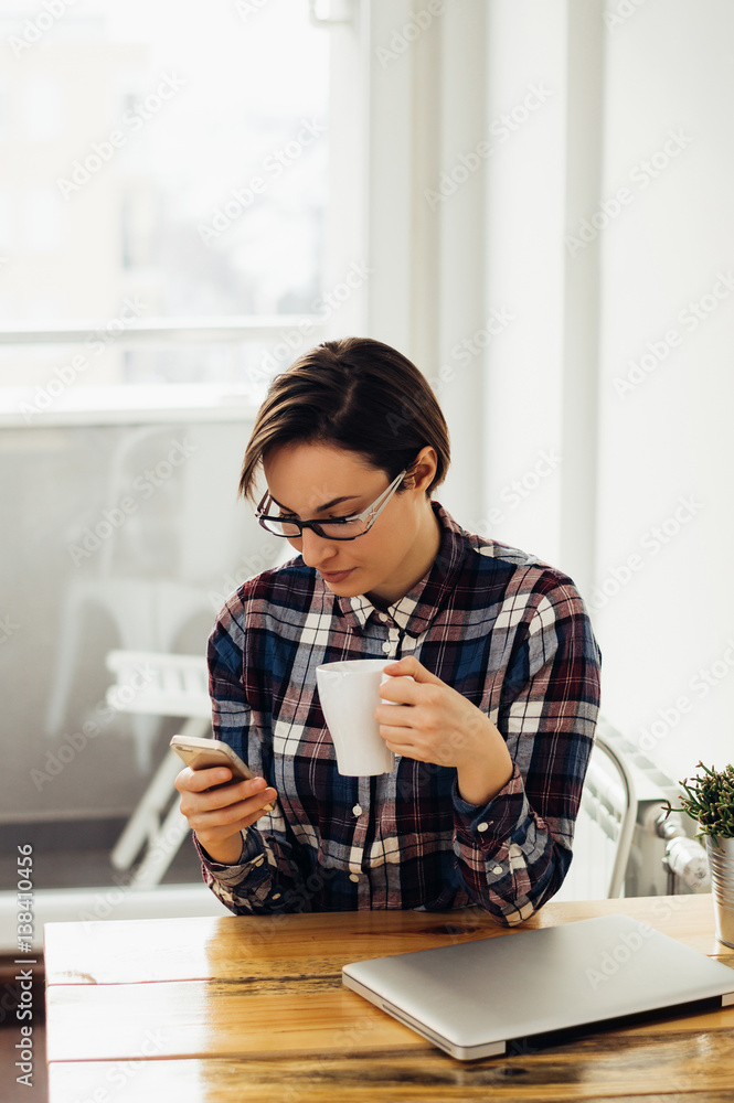 Young woman working in home office. Checking mobile while having a cup of tea or coffee