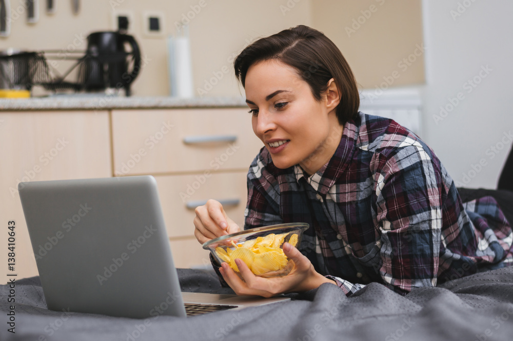 Girl watching a movie on computer and laying on bed, eating chips