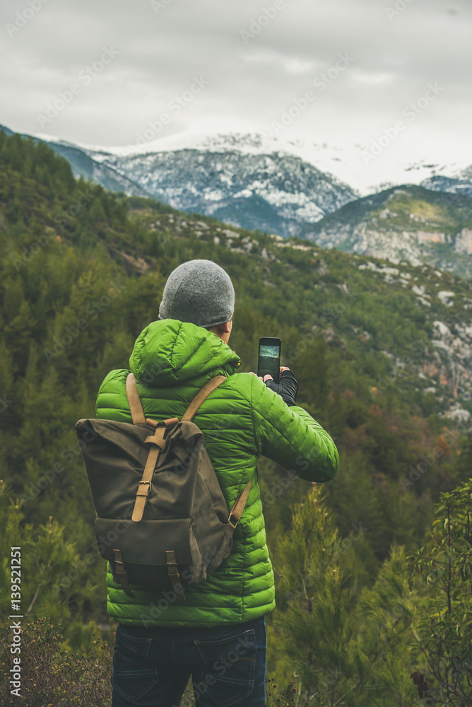 Young man traveller wearing bright jacket making photo of beautiful landscape with slopes and snowy 