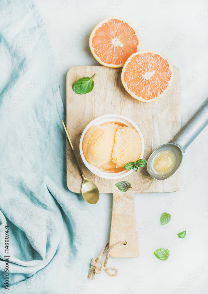 Pink grapefruit homemade sorbet with fresh mint leaves in white bowl on wooden board over light grey