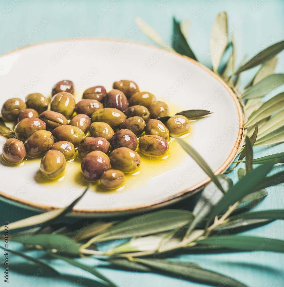 在白色陶瓷盘子和绿色橄榄树枝上用初榨油腌制的绿色地中海橄榄