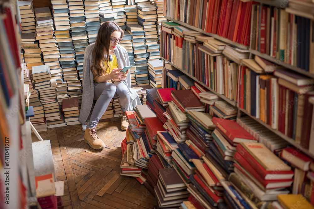 Young woman student sitting on the heap of books at the old library