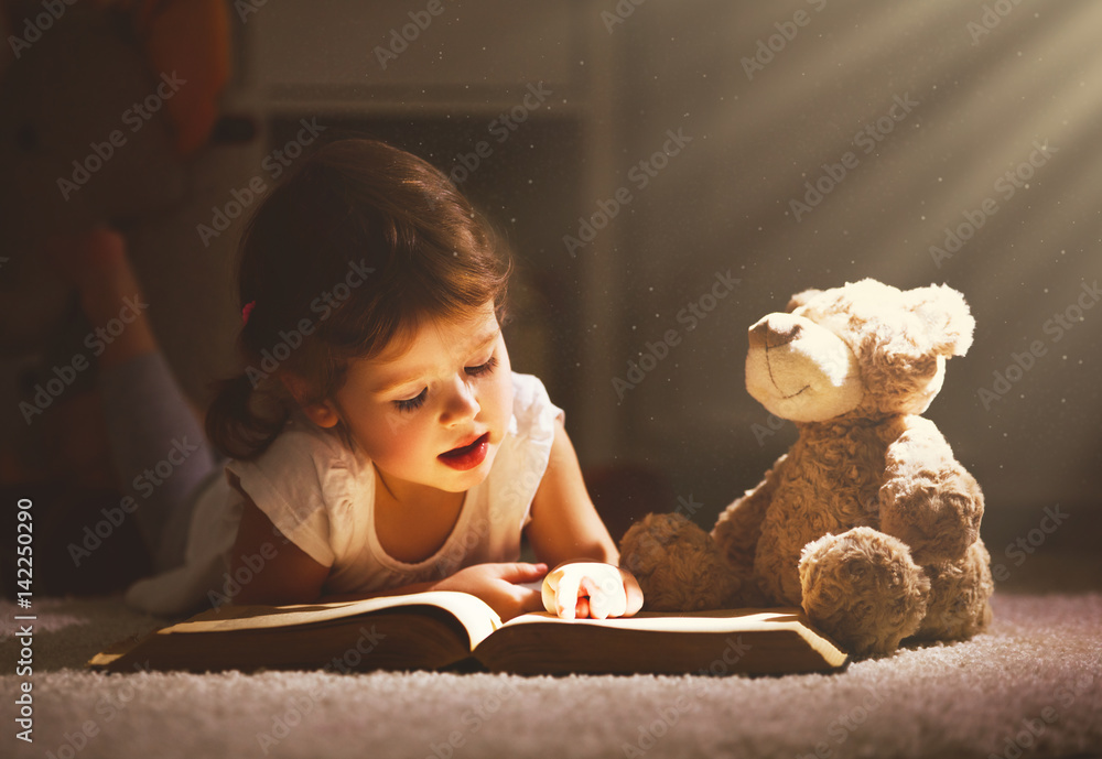一个小女孩晚上在黑暗中和一只玩具熊一起看书。