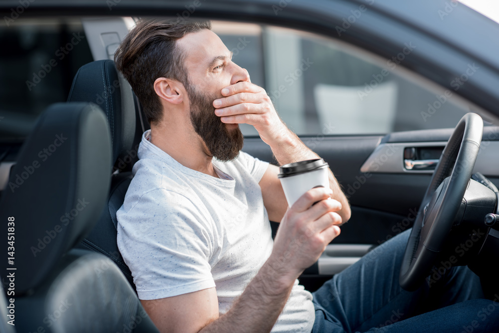一个疲惫的男人在汽车前排座位上打着哈欠，手里拿着咖啡要走