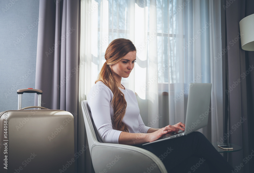现代酒店房间里坐在扶手椅上用笔记本电脑的漂亮女人
