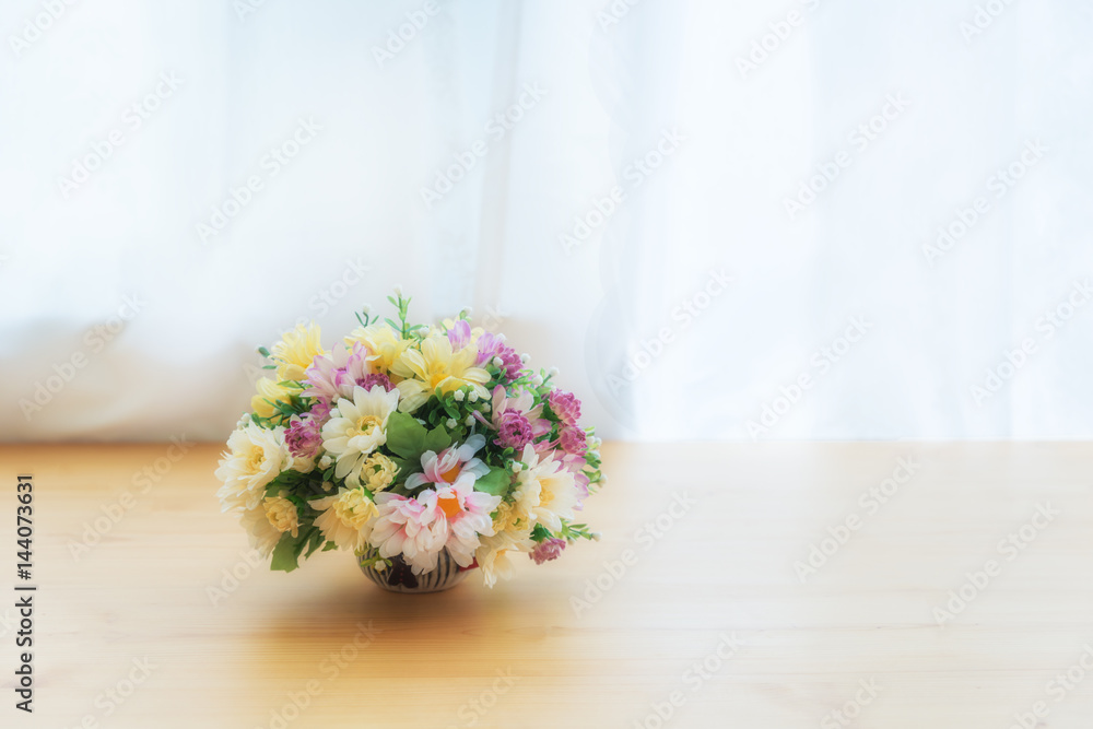 窗帘旁边的木桌子上的陶瓷壶里有五颜六色的人造花。