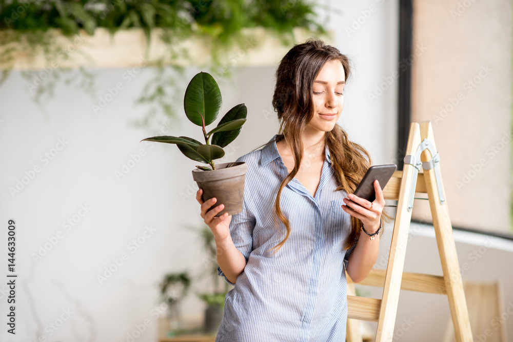 Young woman planting home with greenery standing with phone and flowerpot on the ladder