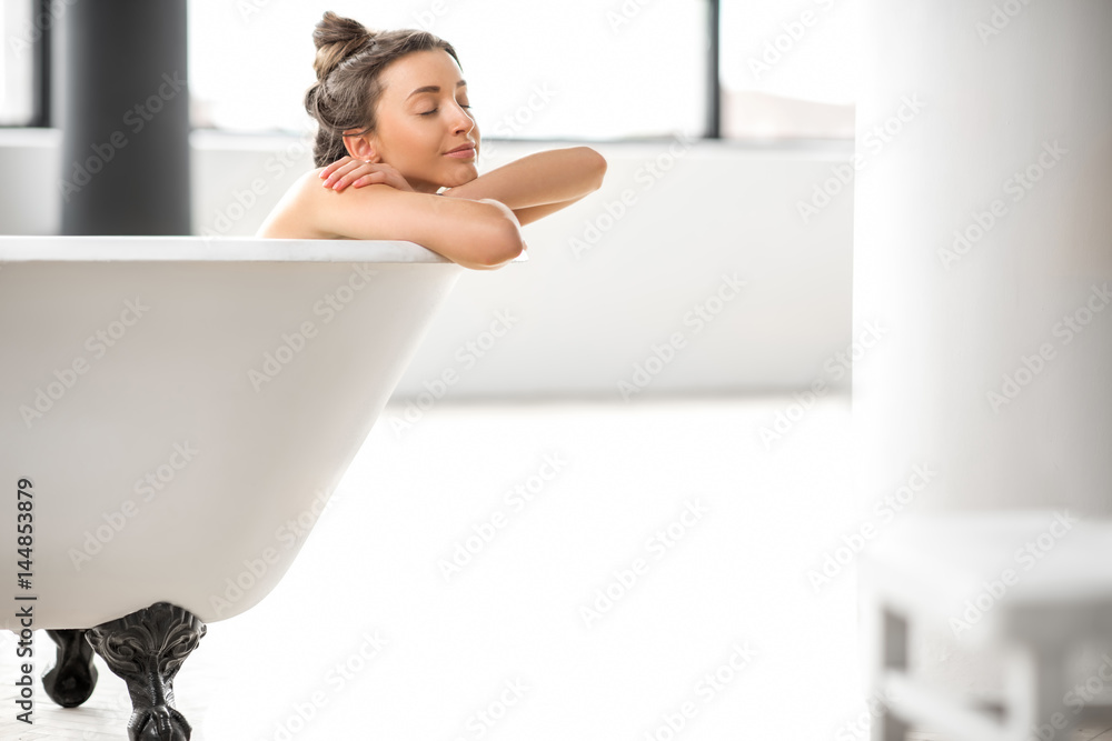 Beautiful young woman relaxing lying in the bathtube at the bright bathroom with windows