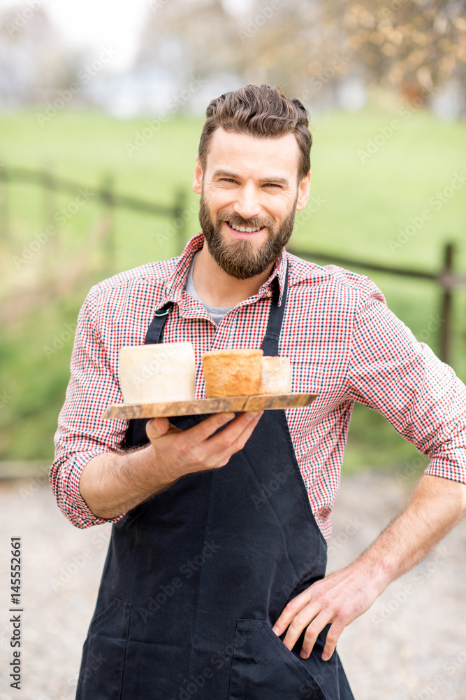 Portrait of a handsome farmer in apron standing with goat cheeses outdoors on the meadow background
