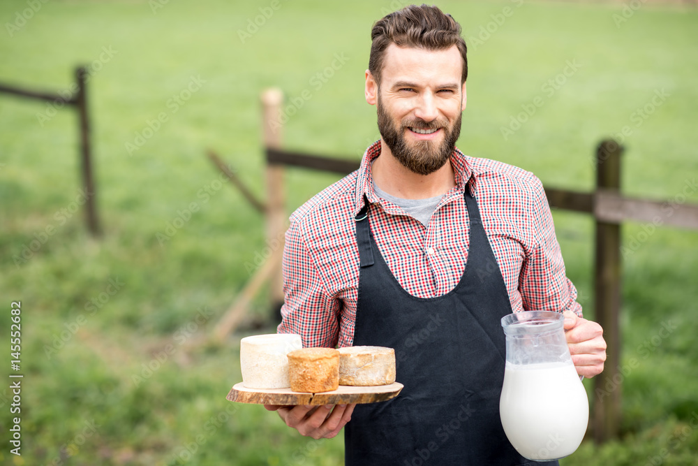 一位英俊的农民穿着围裙，站在户外的绿色美阿上，手里拿着山羊奶酪和牛奶