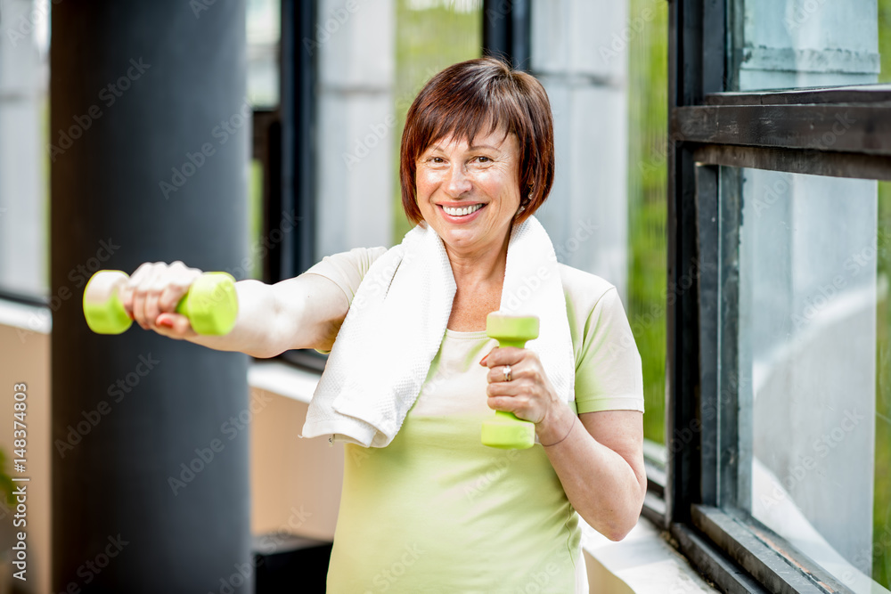 Happy older woman in sports wear training with dumbbells indoors on the window background