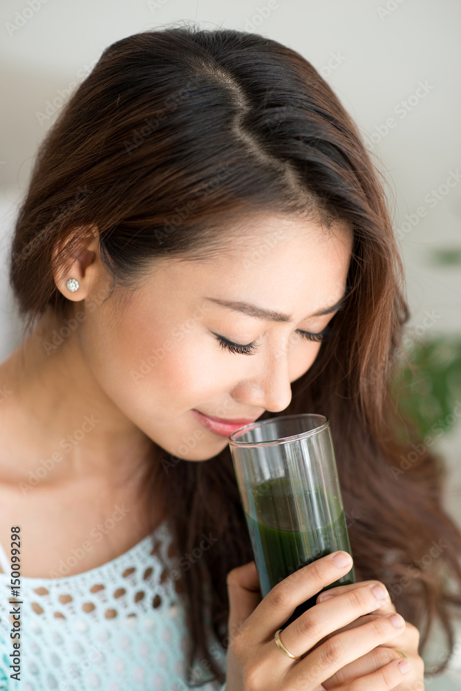 Pretty young asian woman holding green fresh vegetable juice or smoothie from glass at home