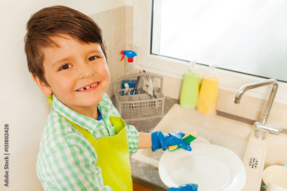 Happy boy washing dishes standing next to the sink
