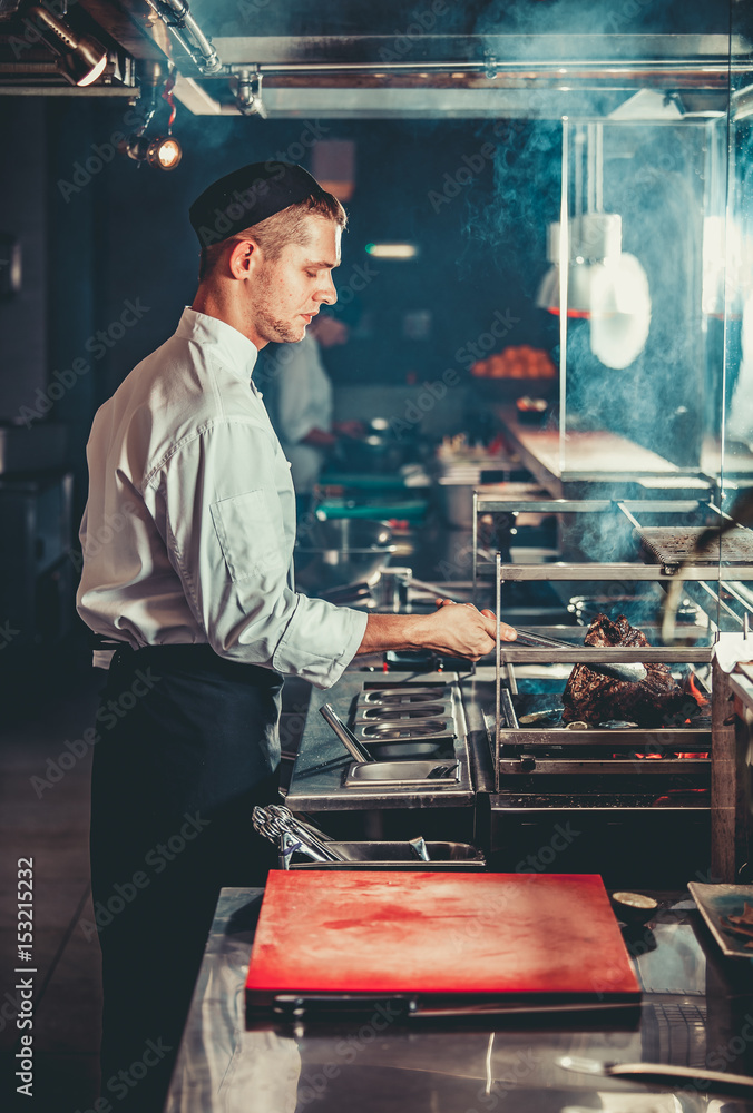 Food concept. Young handsome chef in white uniform monitors the degree of roasting and turns meat wi