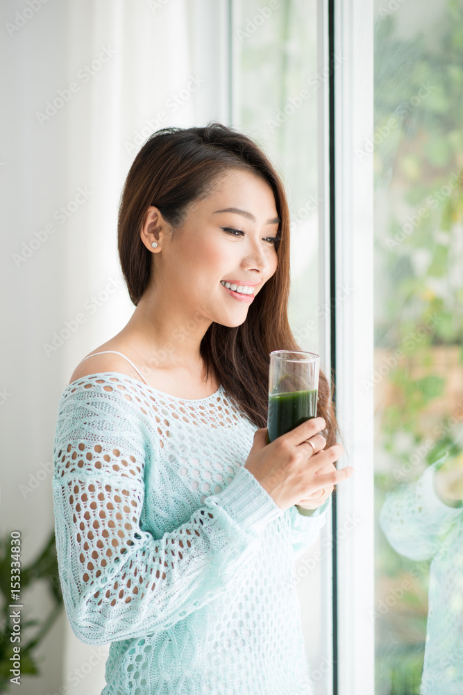 微笑的年轻亚洲女性在家喝绿色新鲜蔬菜汁或冰沙