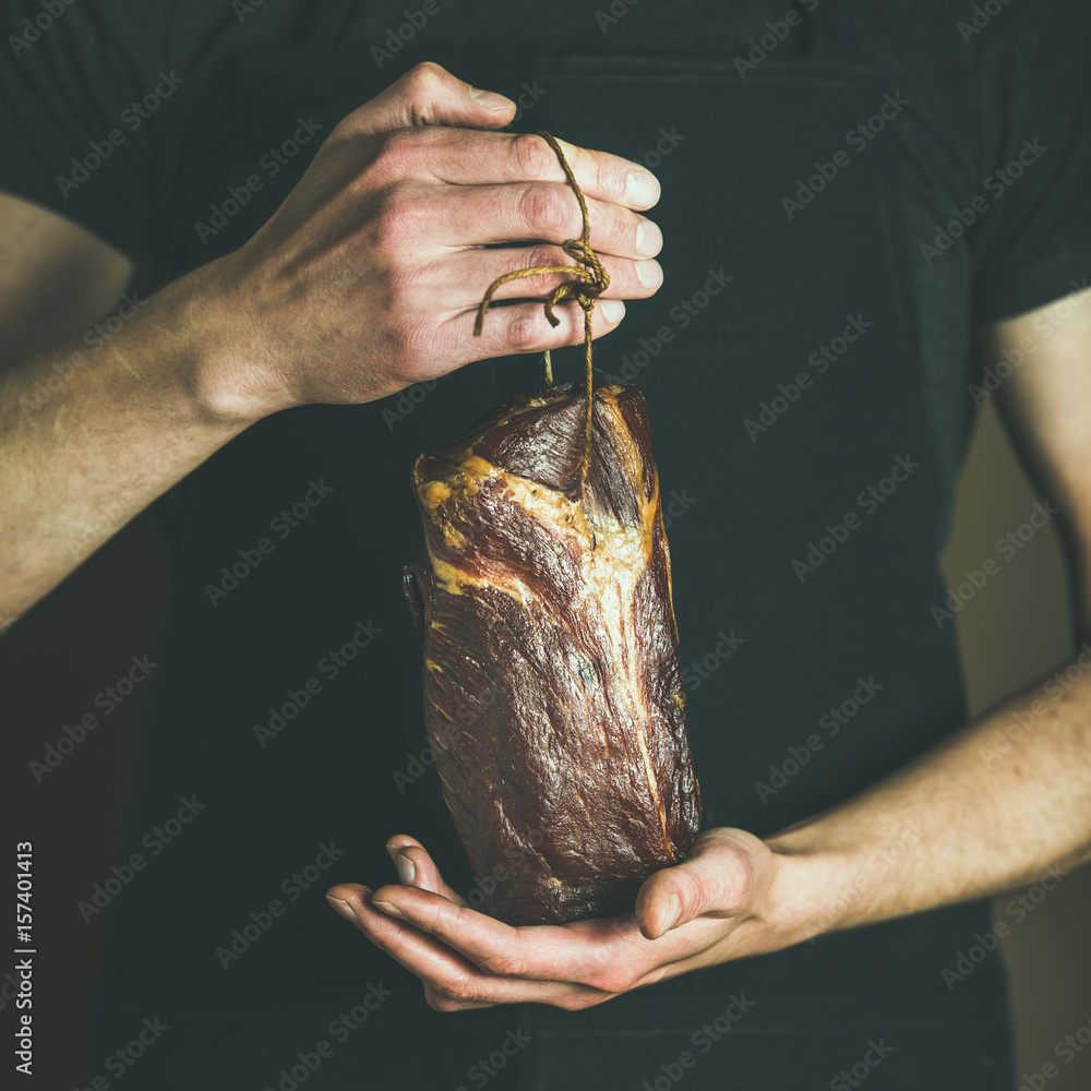 在当地农贸市场，一名身穿黑色围裙的男子手里拿着一块腌猪肉，方形作物。