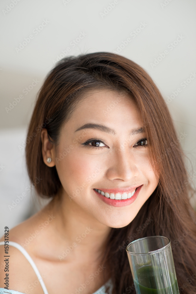 漂亮的年轻亚洲女人在家里拿着杯子里的绿色新鲜蔬菜汁或奶昔